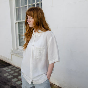 Model wearing white short sleeved loose fit linen shirt with embroidered front pocket detail. Button through front, elbow length sleeves.