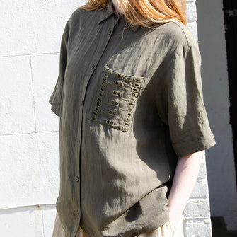 Woman wearing khaki linen short sleeved, loose fitting shirt with embroidery anglaise detail patch pocket to the chest. Button through front.