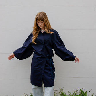 Woman wearing navy blue organic cotton short wrap dress worn as a top over trousers. Full sleeve detailing and adjustable tie front fastening. 