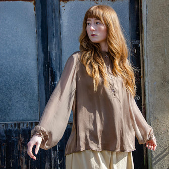 Woman wearing coffee coloured fluid silk shirt with sheer sleeves. Featuring half button front, gathered collar and elastic cuffs.